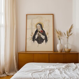 Op de afbeelding: Een ingelijste kunstdruk met een gestileerde illustratie van een figuur in een zwart habijt met een gouden halo, die een houten kruis en een rode roos vasthoudt. Het kunstwerk staat op een cr&egrave;mekleurige achtergrond met korenaren aan weerszijden.