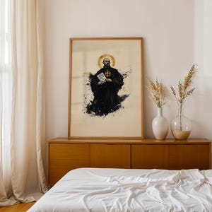 May include: A framed vertical art print featuring a bald monk in a black habit with a gold halo, holding a white document with a cross emblem. The illustration uses black ink splatters and gold accents on a light beige background in a wooden frame.