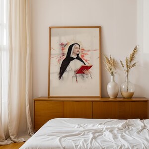 May include: A framed wall art print featuring a portrait of a nun wearing a black and white habit, holding a bright red book. Behind the figure is a vibrant, abstract watercolor-style burst of red and orange light, creating a halo effect.