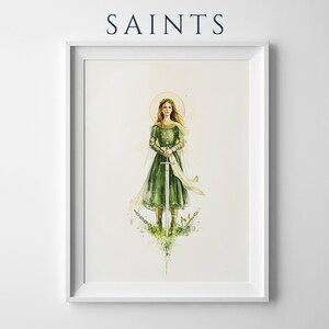 Op de afbeelding: Een ingelijste kunstdruk getiteld SAINTS met een aquarelillustratie van een figuur in een groene jurk die een zwaard vasthoudt. Het figuur heeft een gouden halo, een zwierige sjaal en staat boven gebroken kettingen met groene bladeren.