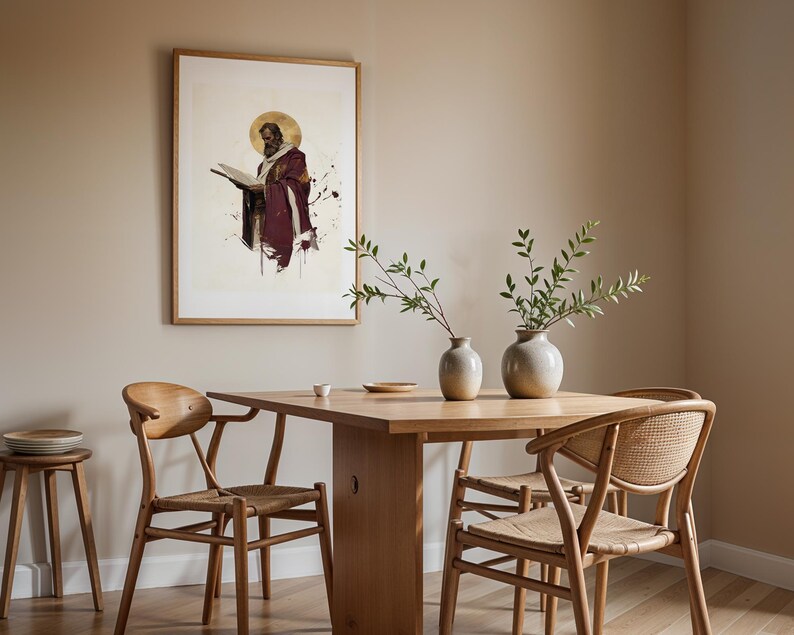 May include: A framed wall art print depicting a bearded figure in a deep red robe holding an open book with a gold halo, displayed above a light wood dining table with four chairs and two ceramic vases containing green leafy branches.