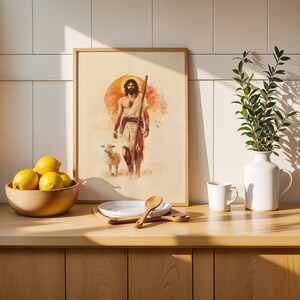 May include: A framed watercolor-style print depicting a bearded figure with a staff walking beside a lamb against a large orange sun. The artwork is displayed on a wooden counter next to a bowl of yellow lemons, a white plate, a spoon, and a white vase.