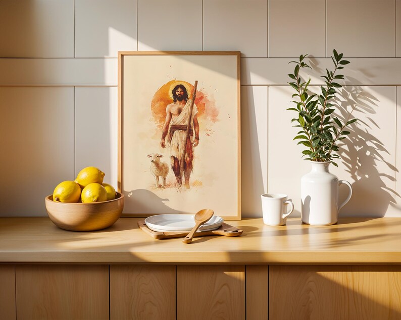 May include: A framed watercolor-style print depicting a bearded figure with a staff walking beside a lamb against a large orange sun. The artwork is displayed on a wooden counter next to a bowl of yellow lemons, a white plate, a spoon, and a white vase.