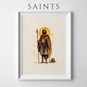 Op de afbeelding: Een ingelijste kunstdruk getiteld SAINTS met een bebaarde figuur in bruine gewaden die een houten staf vasthoudt. De figuur heeft een gouden halo achter het hoofd en staat naast een bruine hoed op een lichte cr&egrave;mekleurige achtergrond met aquarel.