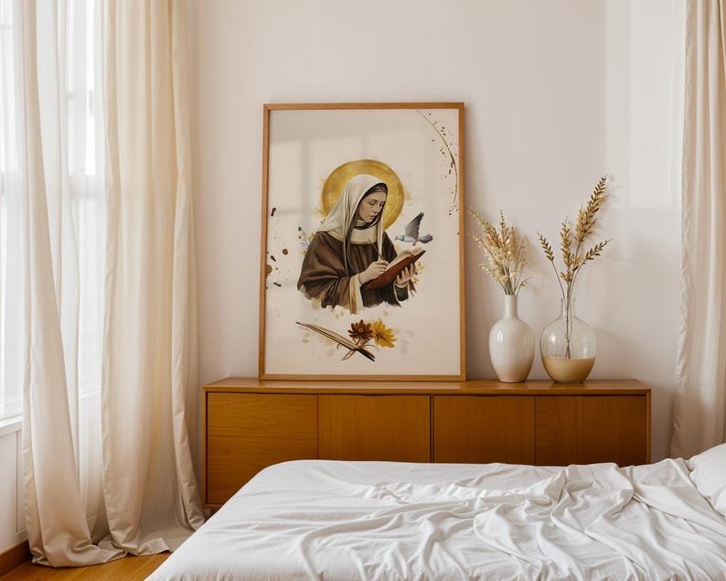 May include: A framed wall art print featuring a figure in a brown habit with a white head covering, writing in a book with a gold halo behind the head. A small bird flies near the figure, with brown and yellow flowers at the bottom of the print.