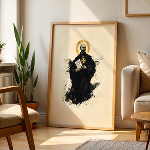 May include: A framed vertical art print featuring a stylised illustration of a bald monk in a black robe with a gold halo. The figure holds a white book with a gold cross emblem. The background is a cream colour with black ink splatter effects.