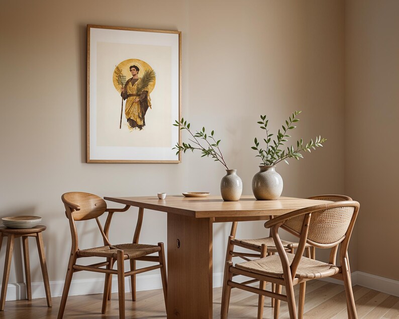 Op de afbeelding: Een ingelijste print van een figuur in gouden gewaden met een palmblad, geplaatst boven een houten eettafel met twee lichtgekleurde keramische vazen met groene takken en drie houten stoelen met gevlochten zittingen.