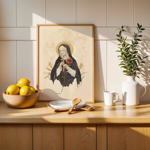 Op de afbeelding: Een ingelijste kunstdruk met een heilige figuur in een zwart habijt die een houten kruis en een rode roos vasthoudt tegen een cr&egrave;mekleurige achtergrond. De illustratie bevat een gouden halo en korenaren, gepresenteerd in een lichte houten lijst.