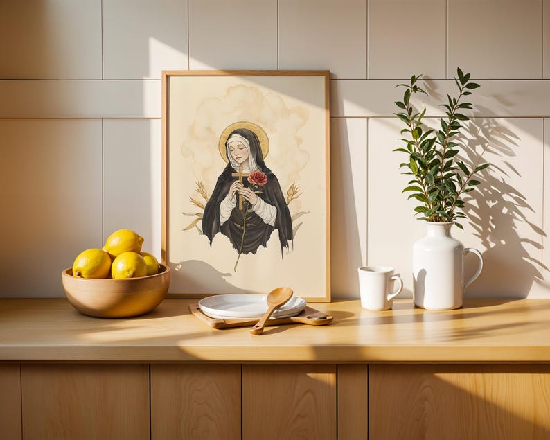 Op de afbeelding: Een ingelijste kunstdruk met een heilige figuur in een zwart habijt die een houten kruis en een rode roos vasthoudt tegen een cr&egrave;mekleurige achtergrond. De illustratie bevat een gouden halo en korenaren, gepresenteerd in een lichte houten lijst.