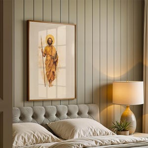 K&ouml;nnte beinhalten: Ein gerahmter Kunstdruck mit einer Aquarell-Illustration einer b&auml;rtigen Figur in goldenen Gew&auml;ndern, die einen langen Stab und ein Buch h&auml;lt, mit einem kreisf&ouml;rmigen Heiligenschein hinter dem Kopf, in einem hellen Holzrahmen an einer Wand.
