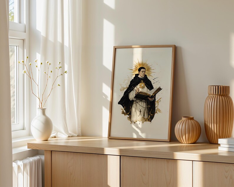 May include: A framed vertical art print featuring a figure in a black and white habit holding an open book, with a golden sunburst halo behind the head. The illustration includes ink-like splatters and a central glowing star emblem on the chest.