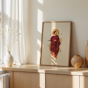 May include: A framed wall art print featuring a watercolour-style illustration of a figure in a deep red robe with a golden halo. The artwork is displayed in a light-toned wooden frame, showcasing a central portrait with abstract paint splatters.