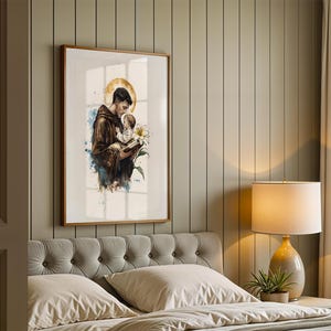 Puede incluir: Una impresi&oacute;n de arte mural vertical enmarcada que presenta una ilustraci&oacute;n en acuarela de una figura con t&uacute;nica marr&oacute;n que sostiene a un ni&ntilde;o peque&ntilde;o y un lirio blanco. Un halo circular dorado se coloca detr&aacute;s de la cabeza de la figura, con salpicaduras de pintura azul y marr&oacute;n.