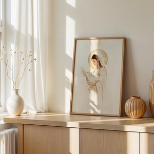 May include: A framed wall art print featuring a stylised illustration of a figure with a golden halo holding a small white lamb and a palm frond. The artwork uses a soft colour palette of cream, white, and gold accents, presented in a light wood frame.