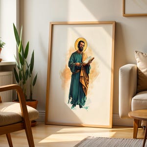 Op de afbeelding: Een ingelijste wandkunstprint met een aquarelillustratie van een bebaard figuur in een blauwgroen gewaad en gouden mantel, die een open boek vasthoudt. Het figuur heeft een gouden halo achter het hoofd tegen een lichte achtergrond.