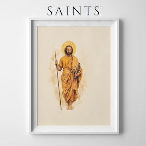 Op de afbeelding: Een ingelijste kunstdruk met de tekst SAINTS bovenaan. Het kunstwerk toont een bebaarde figuur in goudgele gewaden met een aureool, die een lange speer in de rechterhand en een boekrol in de linkerhand houdt, tegen een lichtbeige achtergrond.