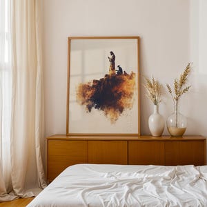 May include: A framed rectangular wall art print featuring a watercolour-style illustration of a figure standing on a cliff edge with a warm brown and orange colour palette. The artwork is displayed in a light wood frame on a wooden sideboard.