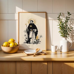 Op de afbeelding: Een ingelijste kunstdruk met een zwart-witillustratie van een figuur in gewaad met een sterrenhalo, een hond en lelies. De print staat op een houten aanrecht naast een kom met gele citroenen, een wit bord en een vaas met groene takken.