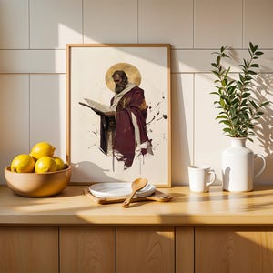 Op de afbeelding: Een ingelijste rechthoekige print met een illustratie van een bebaarde figuur in een bordeauxrode mantel die een open boek vasthoudt, met een gouden halo achter het hoofd. De print staat op een houten aanrecht naast een schaal met gele citroenen.
