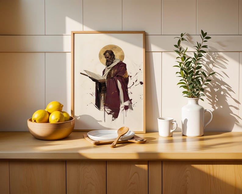 Op de afbeelding: Een ingelijste rechthoekige print met een illustratie van een bebaarde figuur in een bordeauxrode mantel die een open boek vasthoudt, met een gouden halo achter het hoofd. De print staat op een houten aanrecht naast een schaal met gele citroenen.