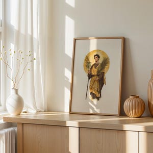 Puede incluir: Una l&aacute;mina enmarcada que muestra una figura con t&uacute;nicas doradas y un halo, sosteniendo una hoja de palma y un bast&oacute;n. La obra est&aacute; en un marco de madera clara, junto a un jarr&oacute;n blanco con ramas finas y dos jarrones de madera acanalados.