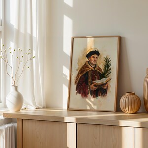 May include: A framed watercolour print depicting a historical figure wearing a dark cap and a fur-trimmed robe, holding an open book and a palm frond. The figure has a golden halo behind their head, set against a light, neutral-toned background.