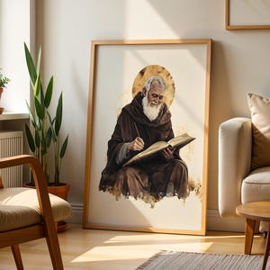 Pode incluir: Uma impress&atilde;o de arte de parede emoldurada apresentando uma ilustra&ccedil;&atilde;o em estilo aquarela de uma pessoa idosa com barba branca vestindo um manto marrom com capuz, sentada e escrevendo em um livro aberto com um efeito de halo dourado.