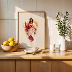 May include: A framed watercolor-style print depicting a figure with a crown of thorns and a glowing red heart on the chest, surrounded by a warm orange and red aura. The artwork is displayed on a counter next to a bowl of lemons and a white vase.