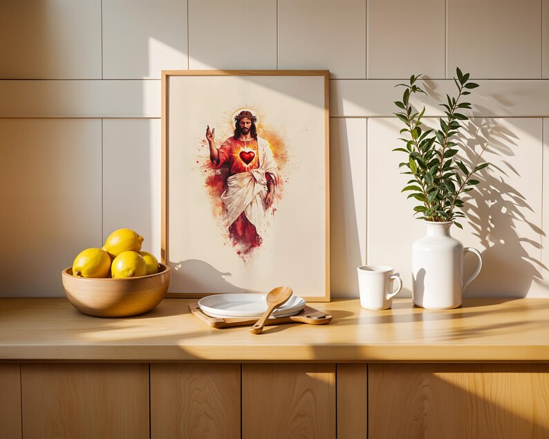 May include: A framed watercolor-style print depicting a figure with a crown of thorns and a glowing red heart on the chest, surrounded by a warm orange and red aura. The artwork is displayed on a counter next to a bowl of lemons and a white vase.