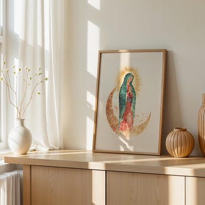 May include: A framed art print featuring a watercolor illustration of the Virgin of Guadalupe standing on a crescent moon, surrounded by a golden sunburst halo. The artwork is displayed in a light wood frame with a cream-colored background.