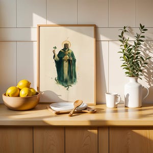 May include: A framed vertical art print featuring a bearded figure in a dark green robe and crown, holding a shamrock and a staff. The illustration is set against a cream background, displayed in a light wood frame on a kitchen counter.