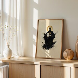 May include: A framed vertical print featuring a stylized illustration of a monk in a black habit with a gold halo, holding a white document. The artwork displays dark ink splatters and is displayed in a light-colored wooden frame on a surface.