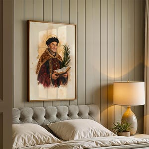 May include: A framed wall art print depicting a historical figure wearing a dark cap and a fur-trimmed robe, holding an open book and a green palm frond. The figure has a golden halo behind their head, rendered in a watercolour style with warm tones.