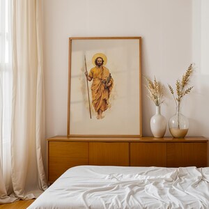 Op de afbeelding: Een ingelijste verticale wandkunstprint met een aquarelillustratie van een bebaard figuur met een gouden halo, gekleed in okerkleurige gewaden, met een lange houten staf in de rechterhand en een boek in de linkerhand.
