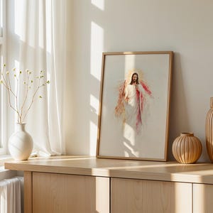 May include: A framed vertical print featuring a watercolor-style illustration of a figure in white robes with a red sash and a glowing golden halo. The artwork is displayed in a light wood frame, positioned alongside a white vase and two wooden vases.