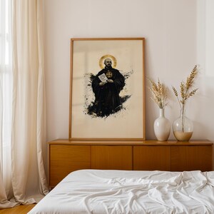 May include: A framed wall art print featuring a bald monk in a black habit with a gold halo, holding a white scroll with text. The artwork uses black ink splatters and gold accents on a light background, presented in a simple wooden frame.
