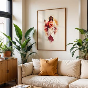 May include: A framed wall art print featuring a watercolor-style illustration of Jesus Christ with a glowing red Sacred Heart, wearing white and red robes, with a golden halo, displayed in a light-colored room with indoor plants and a sofa.