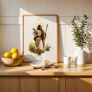 May include: A framed watercolour-style print depicting a figure with long hair and a beard holding a small lamb while carrying a wooden staff. The artwork is displayed on a counter next to a wooden bowl of yellow lemons, a white plate, and a vase.