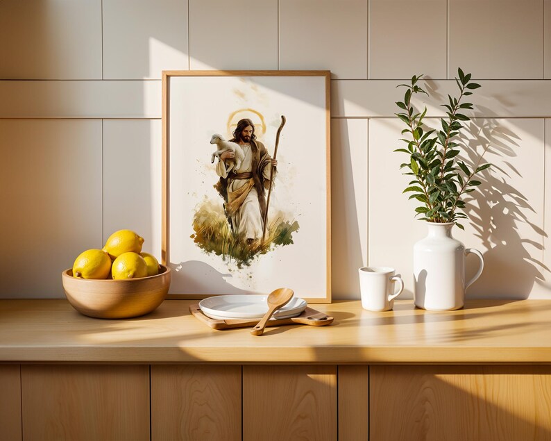 May include: A framed watercolour-style print depicting a figure with long hair and a beard holding a small lamb while carrying a wooden staff. The artwork is displayed on a counter next to a wooden bowl of yellow lemons, a white plate, and a vase.