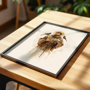 May include: A framed print featuring a watercolor illustration of a figure in a brown robe with a golden halo, holding a white lily, with two small birds perched on the shoulders. The artwork is enclosed in a thin black rectangular frame.