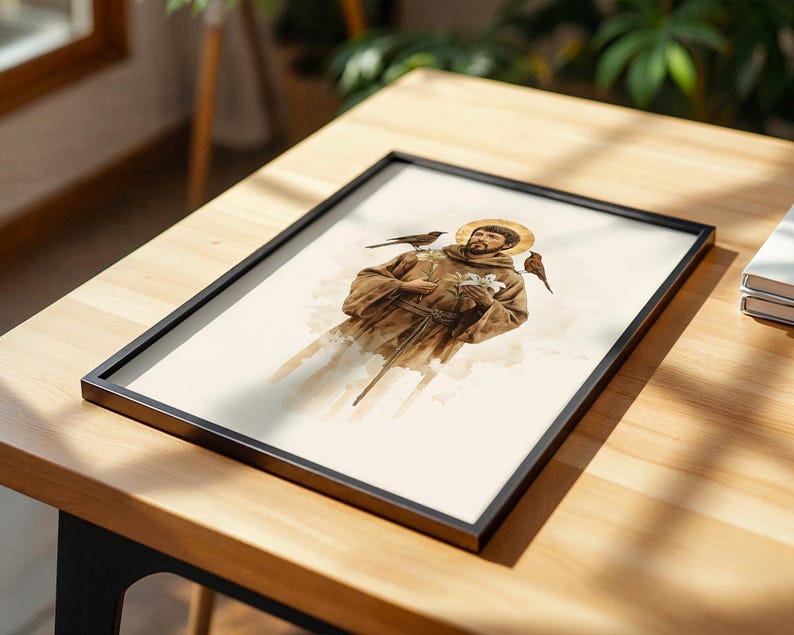 May include: A framed print featuring a watercolor illustration of a figure in a brown robe with a golden halo, holding a white lily, with two small birds perched on the shoulders. The artwork is enclosed in a thin black rectangular frame.