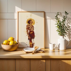 May include: A framed art print featuring a watercolour-style illustration of a person looking upward with a golden halo, displayed on a wooden counter next to a bowl of yellow lemons, a white plate, a wooden spoon, a white mug, and a white vase.