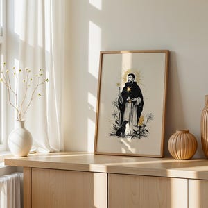 Op de afbeelding: Een ingelijste verticale kunstdruk met een zwart-witillustratie van een figuur in een gewaad met een gouden ster op de borst, omringd door bloemen en een kleine hond, in een lichthouten lijst op een houten ondergrond.