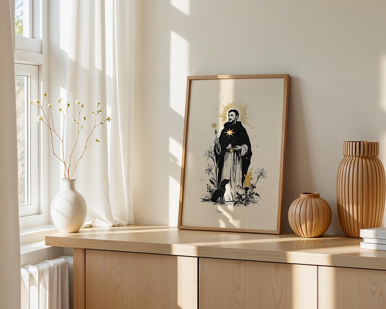 Op de afbeelding: Een ingelijste verticale kunstdruk met een zwart-witillustratie van een figuur in een gewaad met een gouden ster op de borst, omringd door bloemen en een kleine hond, in een lichthouten lijst op een houten ondergrond.