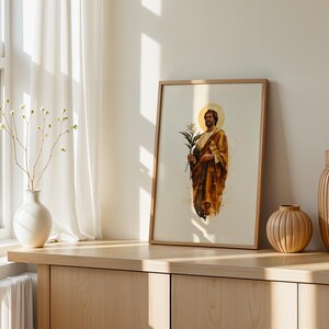 K&ouml;nnte beinhalten: Ein gerahmter vertikaler Druck, der eine b&auml;rtige Figur in einem goldbraunen Gewand mit einem bl&uuml;henden Zweig zeigt, dargestellt mit einem kreisf&ouml;rmigen Heiligenschein hinter dem Kopf auf hellem cremefarbenem Grund in einem hellen Holzrahmen.