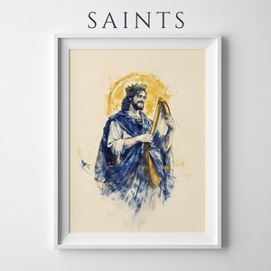 May include: A framed art print titled SAINTS featuring a watercolour-style illustration of a crowned figure with long hair playing a golden harp. The figure wears a flowing blue robe over a white garment, set against a circular yellow halo background.