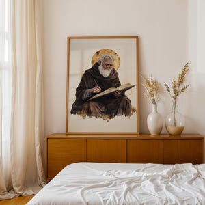 Pode incluir: Uma impress&atilde;o de arte de parede emoldurada com uma ilustra&ccedil;&atilde;o em aguarela de uma pessoa idosa com barba branca vestindo uma t&uacute;nica castanha escura com capuz, escrevendo num livro aberto com um halo dourado atr&aacute;s da cabe&ccedil;a num fundo claro.