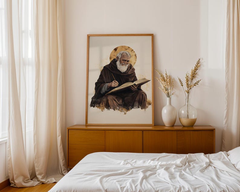 Pode incluir: Uma impress&atilde;o de arte de parede emoldurada com uma ilustra&ccedil;&atilde;o em aguarela de uma pessoa idosa com barba branca vestindo uma t&uacute;nica castanha escura com capuz, escrevendo num livro aberto com um halo dourado atr&aacute;s da cabe&ccedil;a num fundo claro.