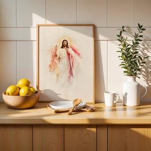 May include: A framed watercolor-style print depicting a figure in white and red robes with a glowing halo, positioned on a wooden counter next to a bowl of yellow lemons, a white ceramic pitcher with green leaves, and a small wooden spoon.