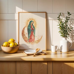 May include: A framed wall art print featuring a watercolor illustration of the Virgin of Guadalupe standing on a crescent moon with a golden sunburst halo, displayed on a wooden counter next to a bowl of yellow lemons and a white ceramic vase.